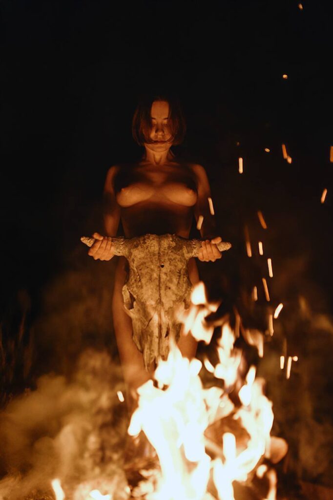 A woman stands before a fire holding a Bison skull, shadow surrounds her bare body illuminated by the flames
