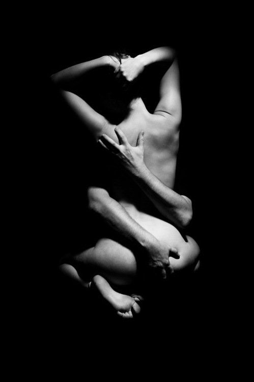 A couple sit in erotic embrace with the man's arms wrapped around his partner, one hand at her sacrum, the other at her shoulder.